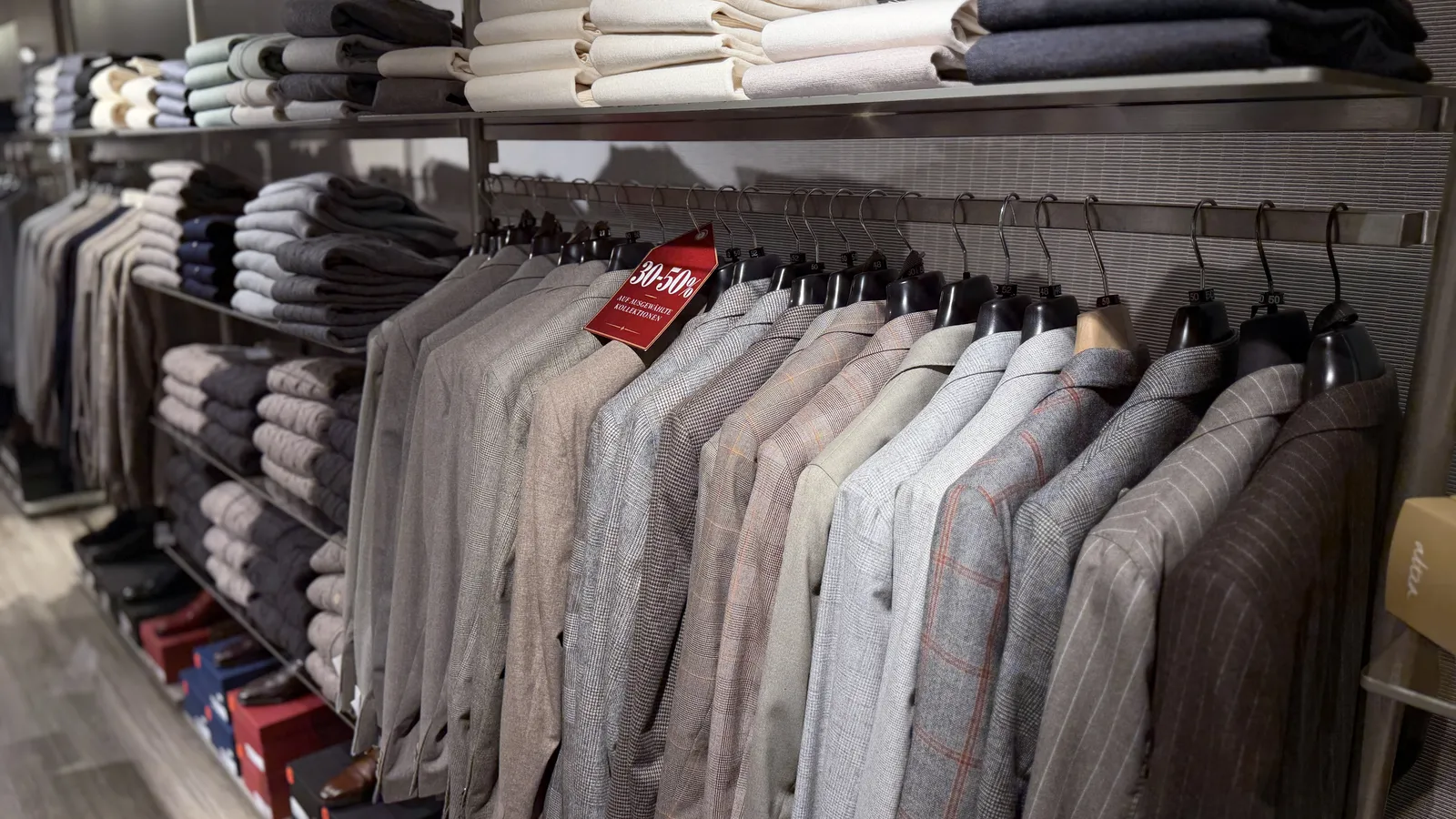 Sakkos in Grau- und Brauntönen an der Wandstange mit Schild „30–50 % auf ausgewählte Kollektionen“, darüber gestapelte Pullover
